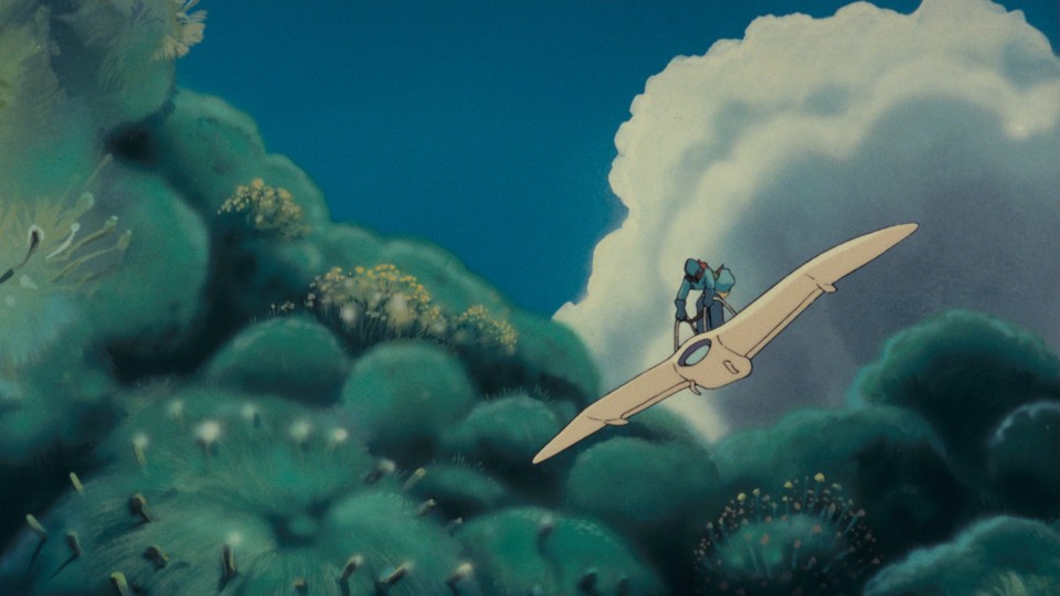 Nausicaä, a small figure in heavy protective clothes, helmet and mask, flies a white albatross-shaped glider over alien-looking plants.