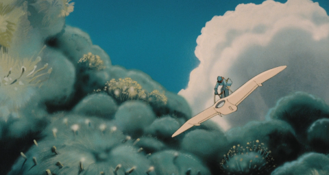 Nausicaä, a small figure in heavy protective clothes, helmet and mask, flies a white albatross-shaped glider over alien-looking plants.