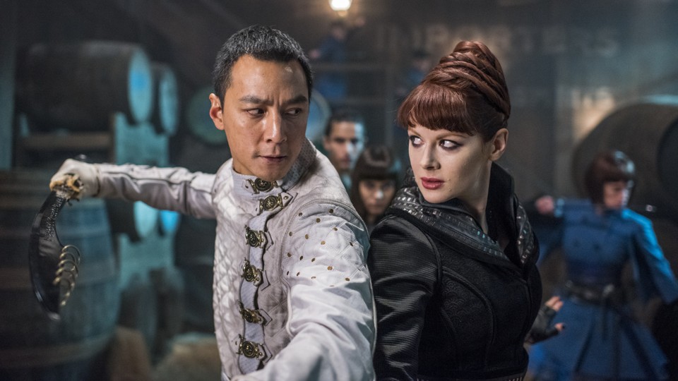 Two figures – an Asian man in a white and a white woman in black – stand shoulder to shoulder, ready to fight.