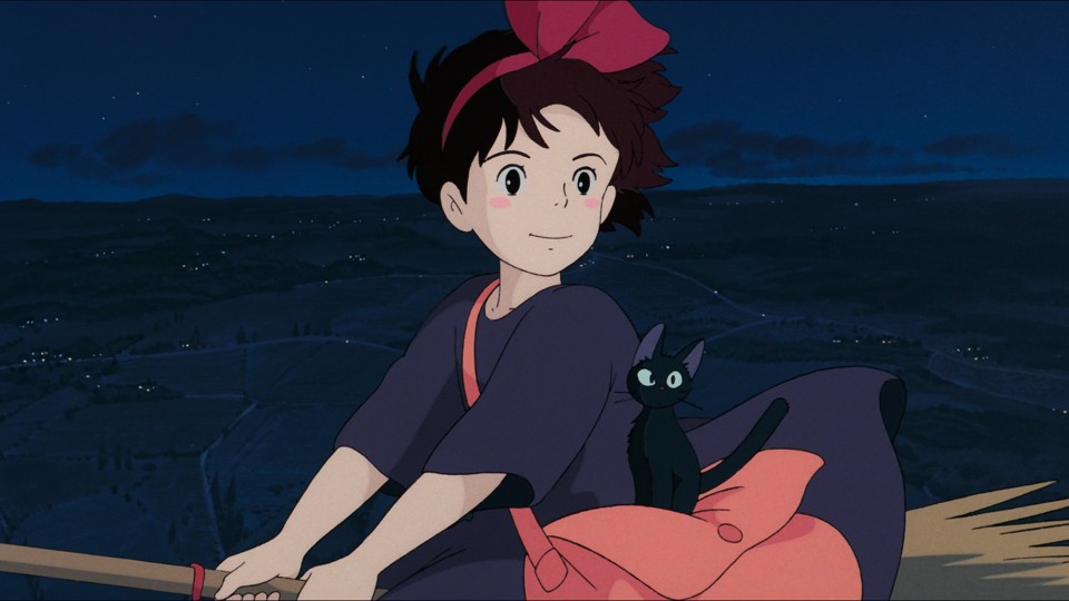 Kiki in a black dress with Jiji, her black cat, on a broom floats in the air over night fields dotted with the lights of small houses.