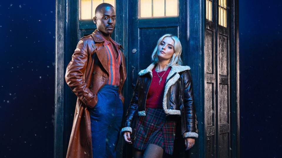 The Doctor, a black man in a long leather coat and blue trousers, and the Doctor's companion, Ruby Sunday, a white blonde woman in a short coat with white fur trim and a dark red tartan skirt, leaning against the TARDIS.