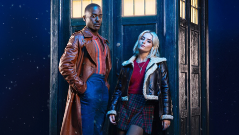 The Doctor, a black man in a long leather coat and blue trousers, and the Doctor&rsquo;s companion, Ruby Sunday, a white blonde woman in a short coat with white fur trim and a dark red tartan skirt, leaning against the TARDIS.