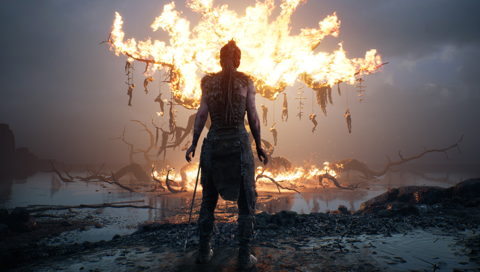 Senua, dressed in leather pants, skirt, and corset, with a sword at her belt, watches the huge burning tree with corpses in tortured poses hanging from its branches.