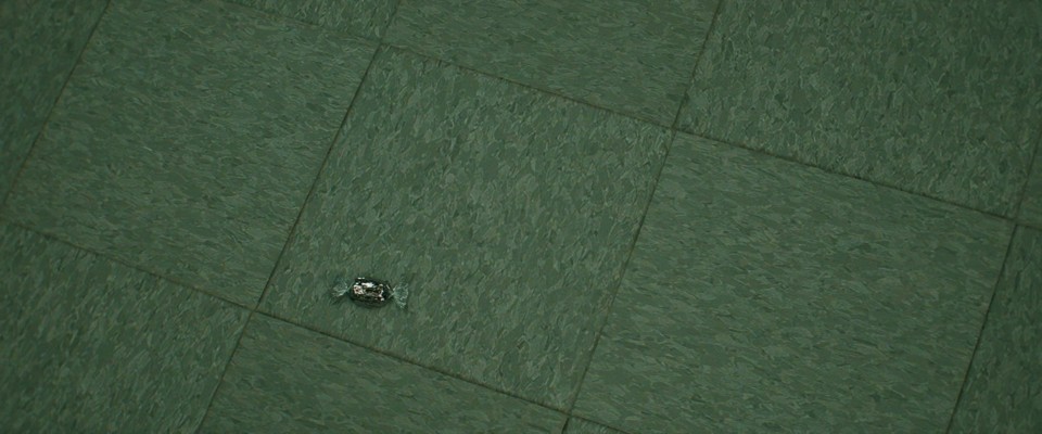 A small piece of hard candy in a plastic wrapper is lying on the gray, tiled floor. A small piece of hard candy in a plastic wrapper is lying on the gray, tiled floor.