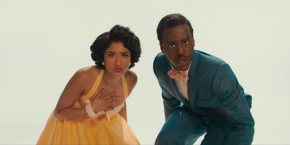 A dark-skinned woman with puffy, curly hair wearing a yellow dress and a dark-skinned man with short hair and a mustache wearing a blue suit with a bow tie. They both leaned over and look intently at the viewer. A dark-skinned woman with puffy, curly hair wearing a yellow dress and a dark-skinned man with short hair and a mustache wearing a blue suit with a bow tie. They both leaned over and look intently at the viewer.