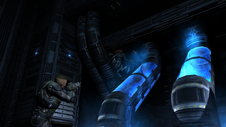 The dark room with metal walls and some pipes, two of which on the left have glass walls that reveal blueish coolant liquid inside. The protagonist, a white man with a short, military-style haircut, wearing dark, metal, camouflage armor, is pointing a machinegun up at an enemy. The enemiy, a cyborg with a humanoid torso and spider-like legs, a head with one glowing eye, is emerging from behind some pipes high up on the wall.