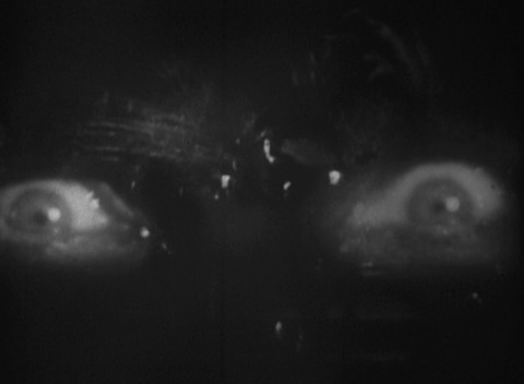 Intense eyes stare at the viewer over a shot of a horse-drawn carriage driving through the woods at night. The image is dark, blurry, and grainy with few details.