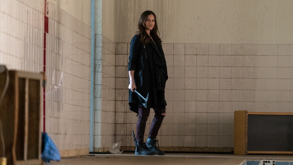 A woman stands in a dirty pool with white tiles covered in grime. There is some wooden furniture and trash on the concrete floor. She is white with long, dark hair and a hint of a “shit-eating grin” on her face. She wears all black, from her long coat to her pants with holes in the knees to her boots. There is a knife in a sheath on her boot, and she is holding a black, full-metal hatchet.