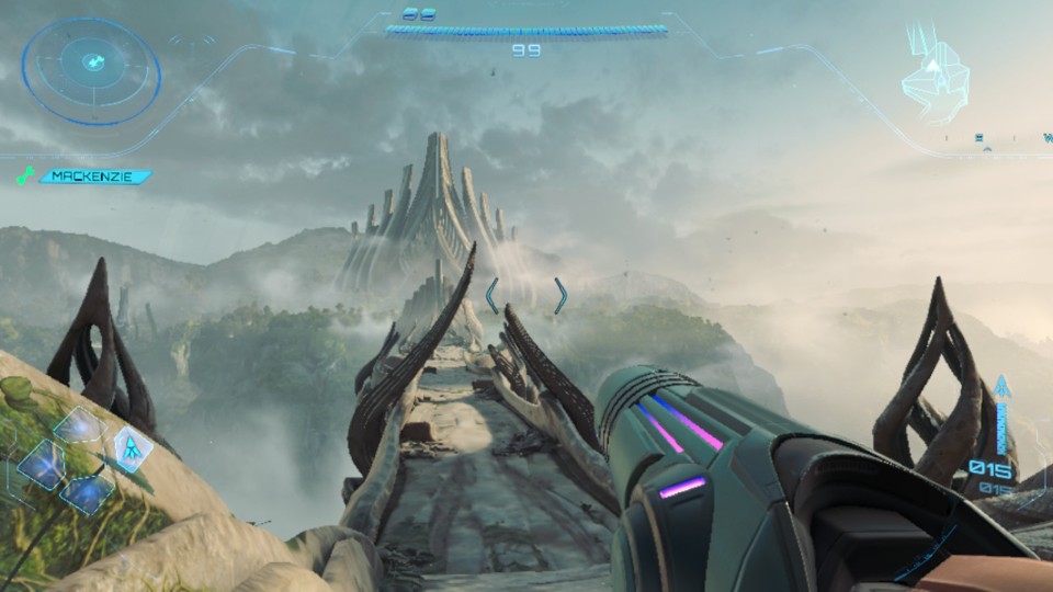 First-person view from Samus’s helmet with the Arm Cannon visible and some usual UI elements, such as mini map, health bar, etc. There is also a name MacKenzie, one of the game’s NPCs, with a wrench icon on the left side. In the background, we see a lush jungle. In the foreground, there is a dilapidated bridge over a canyon that leads to somewhat organic-looking ruins.