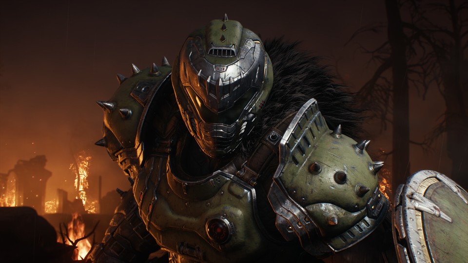 A close-up of the Doom Slayer in his green armor facing the camera. Hints of his facial features are visible behind the dark, V-shaped visor. This time, his armor has a more medieval look: there are spikes on his shoulders and helmet, and corner of the shield is visible. The dark sky behind him is slightly illuminated by the burning ruins on the left and there are a couple of leafless trees on the right.