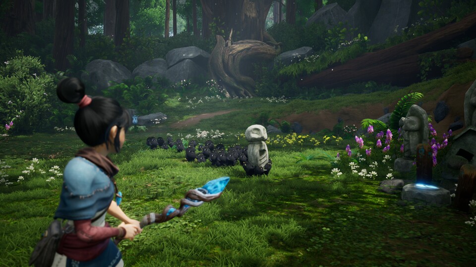 Kena, an young woman with black hair tied in a bun, facing away from the camera. She wears a light sleeveless shirt, her right arm is covered by a red sleeve, and her right shoulder is covered by a short blue cape, almost a pauldron. In her left hand, she holds a staff: a twisted wooden stick with a large crystal. She commands a large group of small, fuzzy, round, black creatures who carry a waist-high stone statue depicting a simple idol to a place near similar statues. The scene is set in a lush forest meadow with big rocks in the background and plenty of flowers.