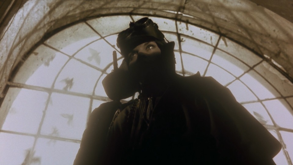 Silhouette of a woman wearing a military-style helmet and metal goggles. Only her upper face, mostly her eyes, is highlighted by a beam of light. A bright sky visible through an arched window in a concrete wall contrasts her. Pigeons fly by.